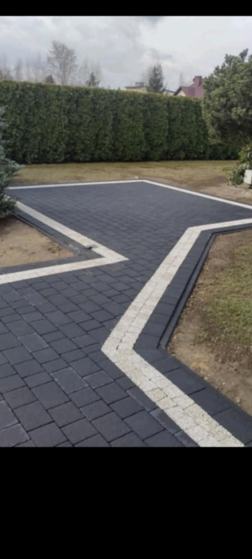 Ułożony podjazd z ciemnoszarej kostki brukowej z dekoracyjnym obramowaniem z jasnej kostki, otoczony trawnikiem i żywopłotem.