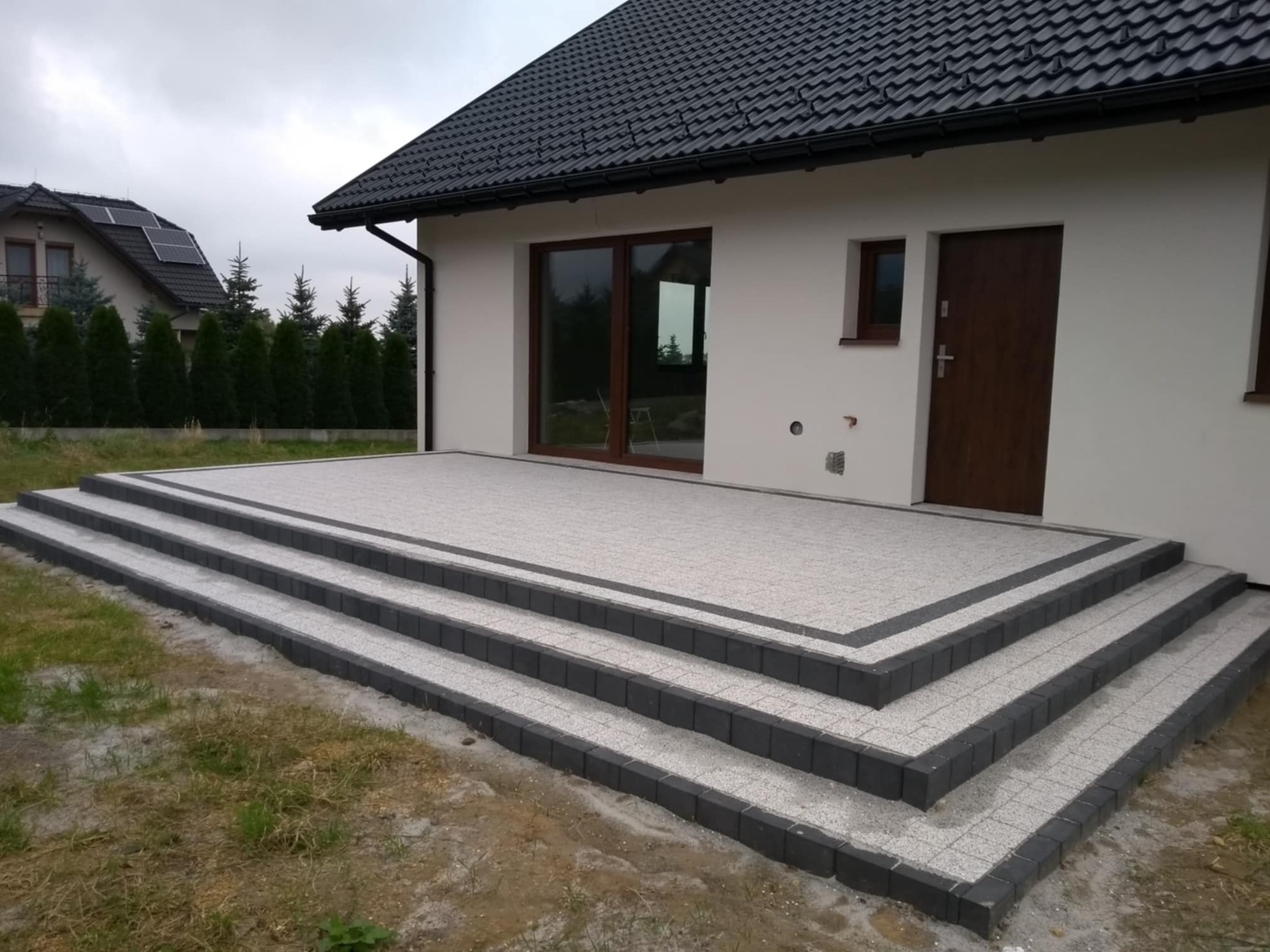 Schody wejściowe do domu wyłożone jasnoszarą kostką brukową z ciemnoszarą obwódką, widok z dołu pod kątem, trawnik w tle.