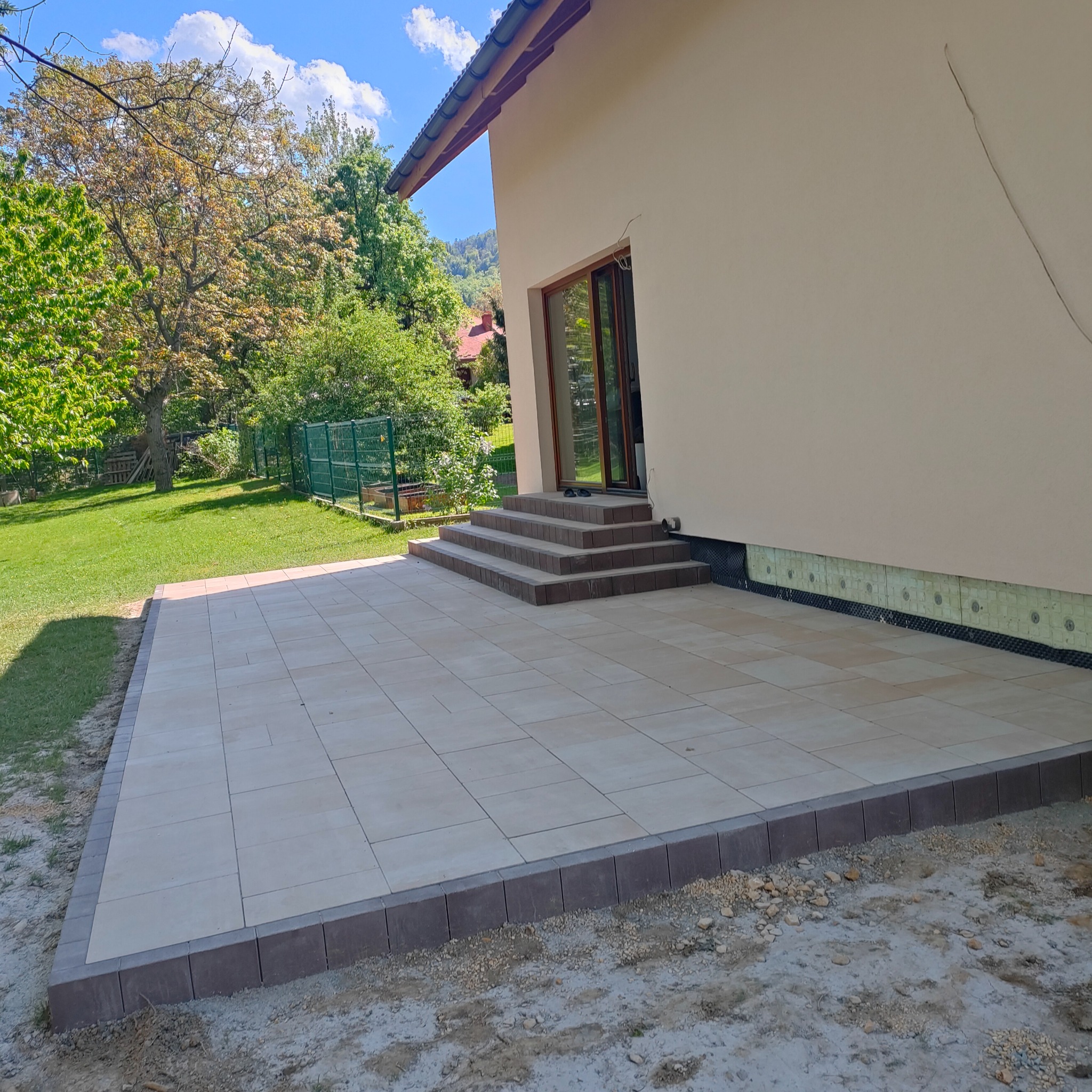 Jasne, prostokątne płyty tarasowe z ciemniejszym obramowaniem, przylegające do beżowego domu z brązowymi schodami i zielonym ogrodem w tle.