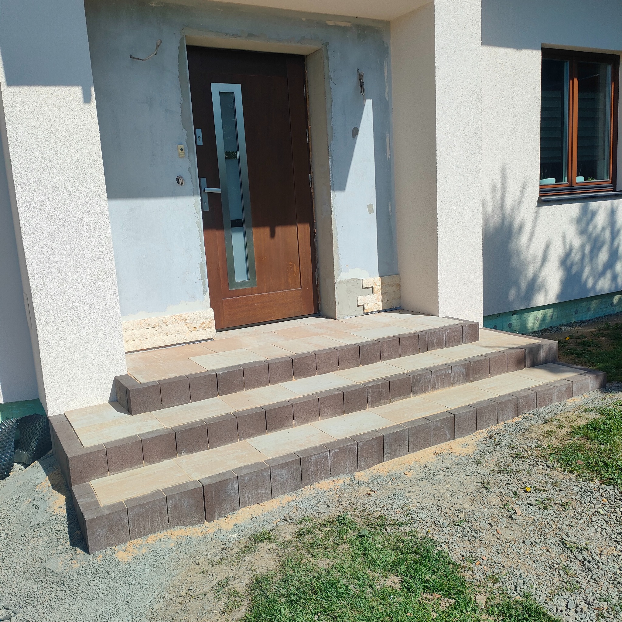 Nowe, trzystopniowe schody z kostki brukowej prowadzą do drzwi wejściowych domu. Kostka dwukolorowa, beżowo-brązowa. Otoczenie w trakcie wykończenia.