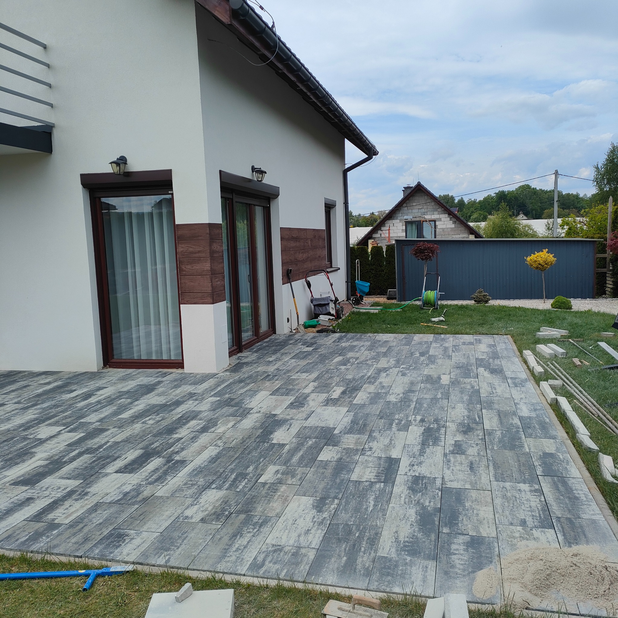 Ułożona kostka brukowa w odcieniach szarości na posesji. Widoczne elementy wykończeniowe, trawnik i fragment budynku z brązowymi oknami. Prace brukarskie w toku.