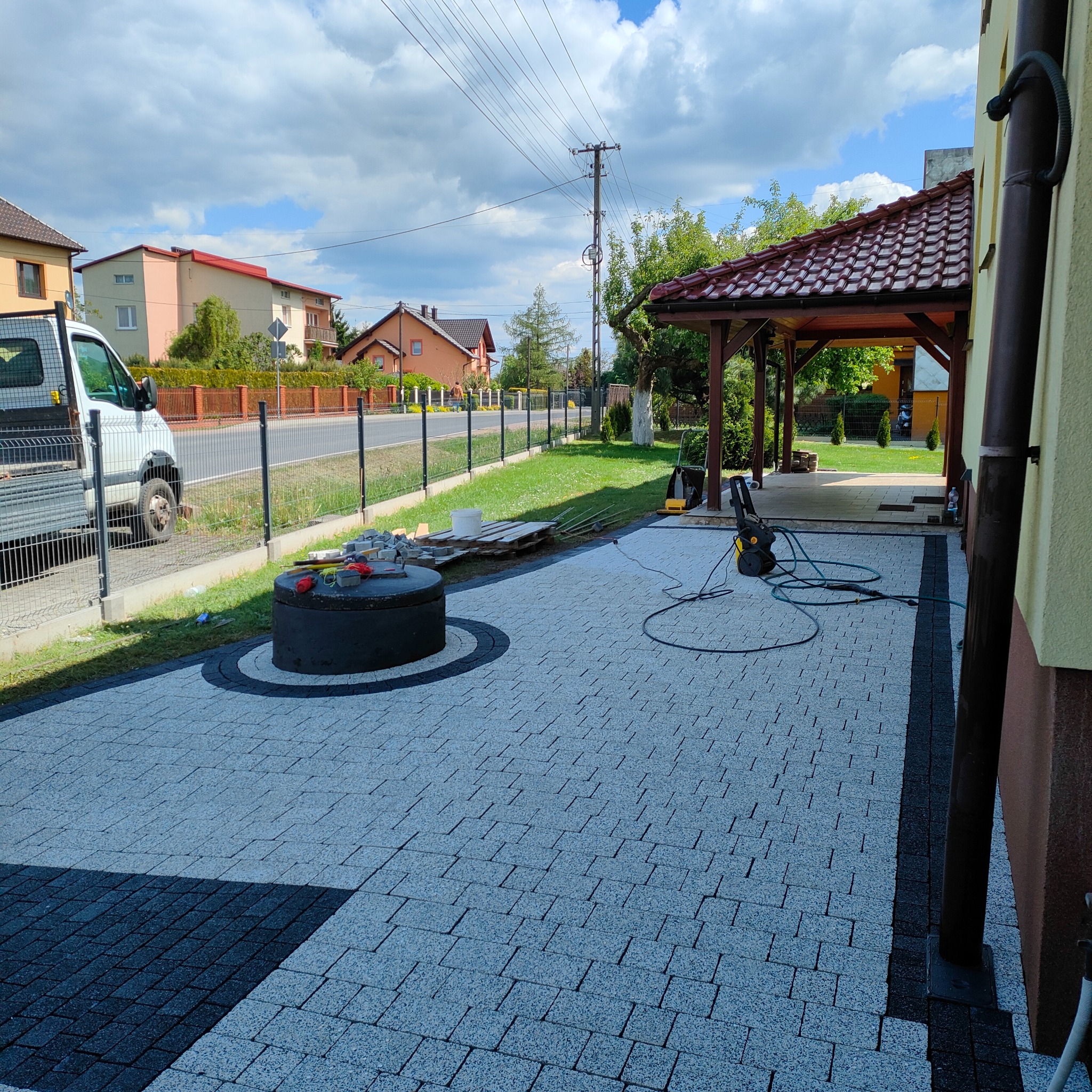 Ułożona kostka brukowa w odcieniach szarości i czerni tworzy wzór na podjeździe. Widoczna studzienka, narzędzia pracy i częściowo biały samochód dostawczy.