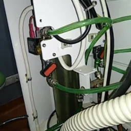 Wnętrze pralki z widocznymi przewodami, zaworami i elementami elektrycznymi, w tym cewką z czerwonymi konektorami, oraz fragmentem zielonego węża doprowadzającego wodę.