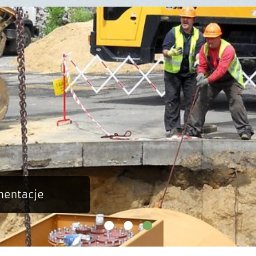 Zmywarka Plus 500 - Pracownicy w kaskach i kamizelkach odblaskowych nad wykopem, ciężki sprzęt budowlany w tle, zabezpieczenie taśmą ostrzegawczą, widoczny fragment zbiornika w wykopie.