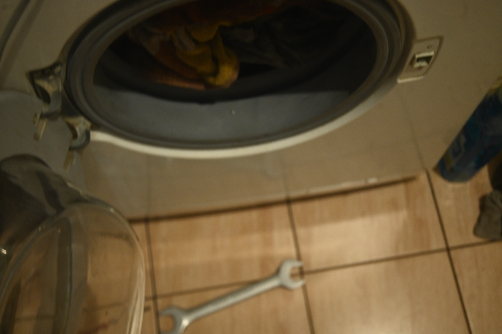 Otwarty bęben pralki wypełniony częściowo ubraniami, z widocznym kluczem płaskim leżącym na brązowych płytkach podłogowych i butelką płynu do prania obok, sugerujący trwające prace serwisowe.