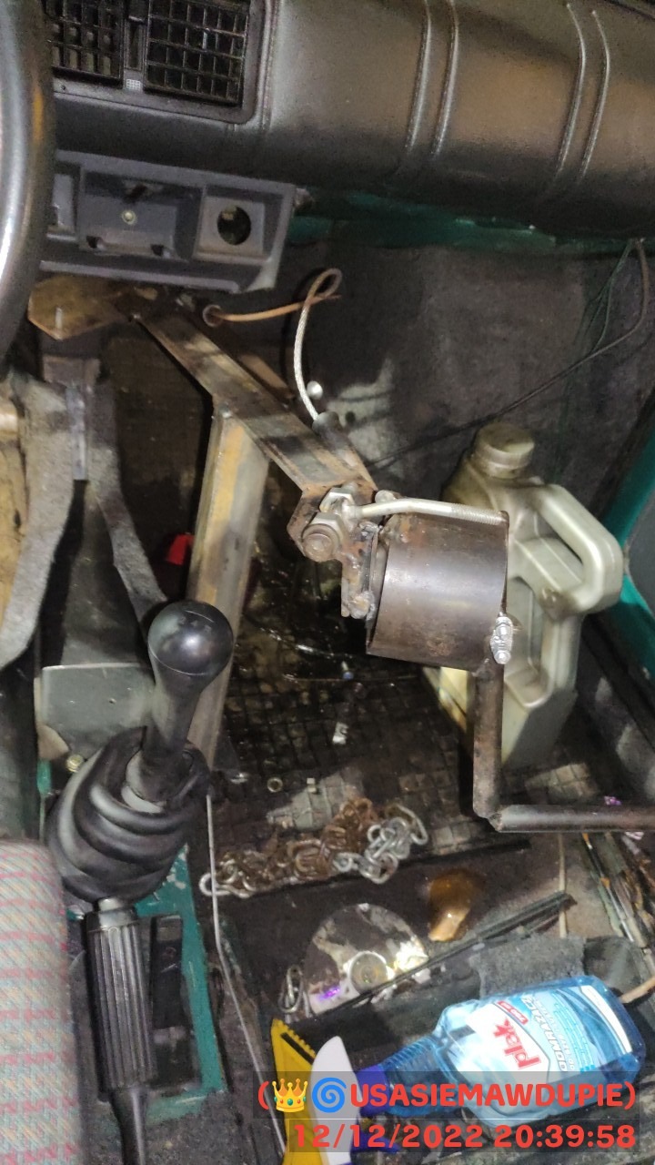 Wnętrze starego, zaniedbanego samochodu z widoczną dźwignią zmiany biegów, metalową konstrukcją, butelką płynu do czyszczenia i łańcuchem na podłodze. Widoczny fragment deski rozdzielczej i tunelu...