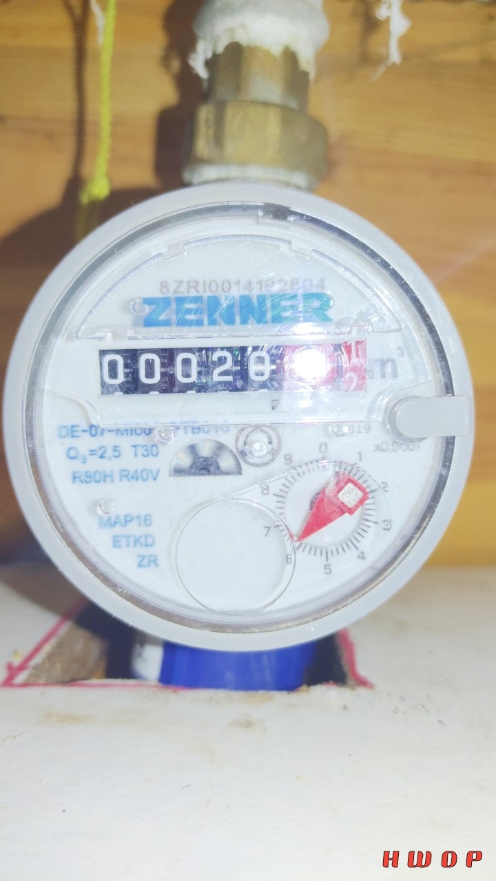 Licznik wody Zenner z widocznym wskazaniem 0.0020 m3, zamontowany na rurze, z uszczelnieniem na gwincie i drewnianym tłem.