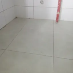 Świeżo ułożona, jasnoszara podłoga z płytek w rogu pomieszczenia, widoczna poziomica oparta o ścianę i wystające rury instalacji wodnej.