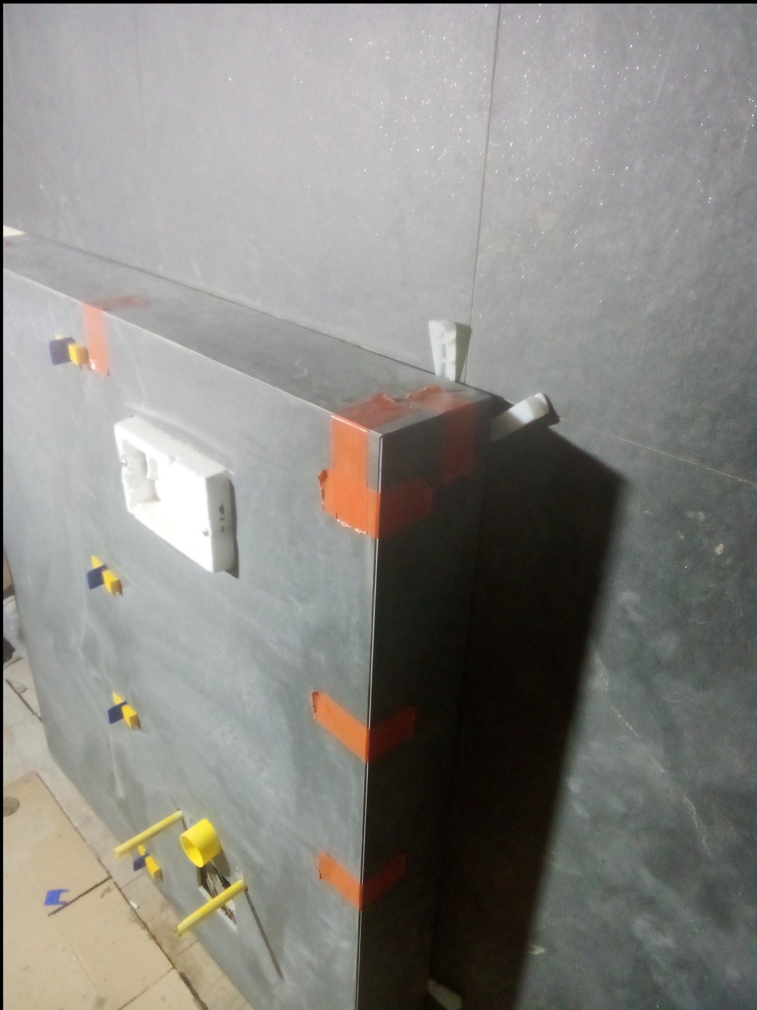 Szare płytki ścienne z pomarańczową taśmą maskującą na krawędziach, żółtymi i niebieskimi krzyżykami dystansowymi oraz białą puszką elektryczną podtynkową, widoczne elementy instalacji wodnej.