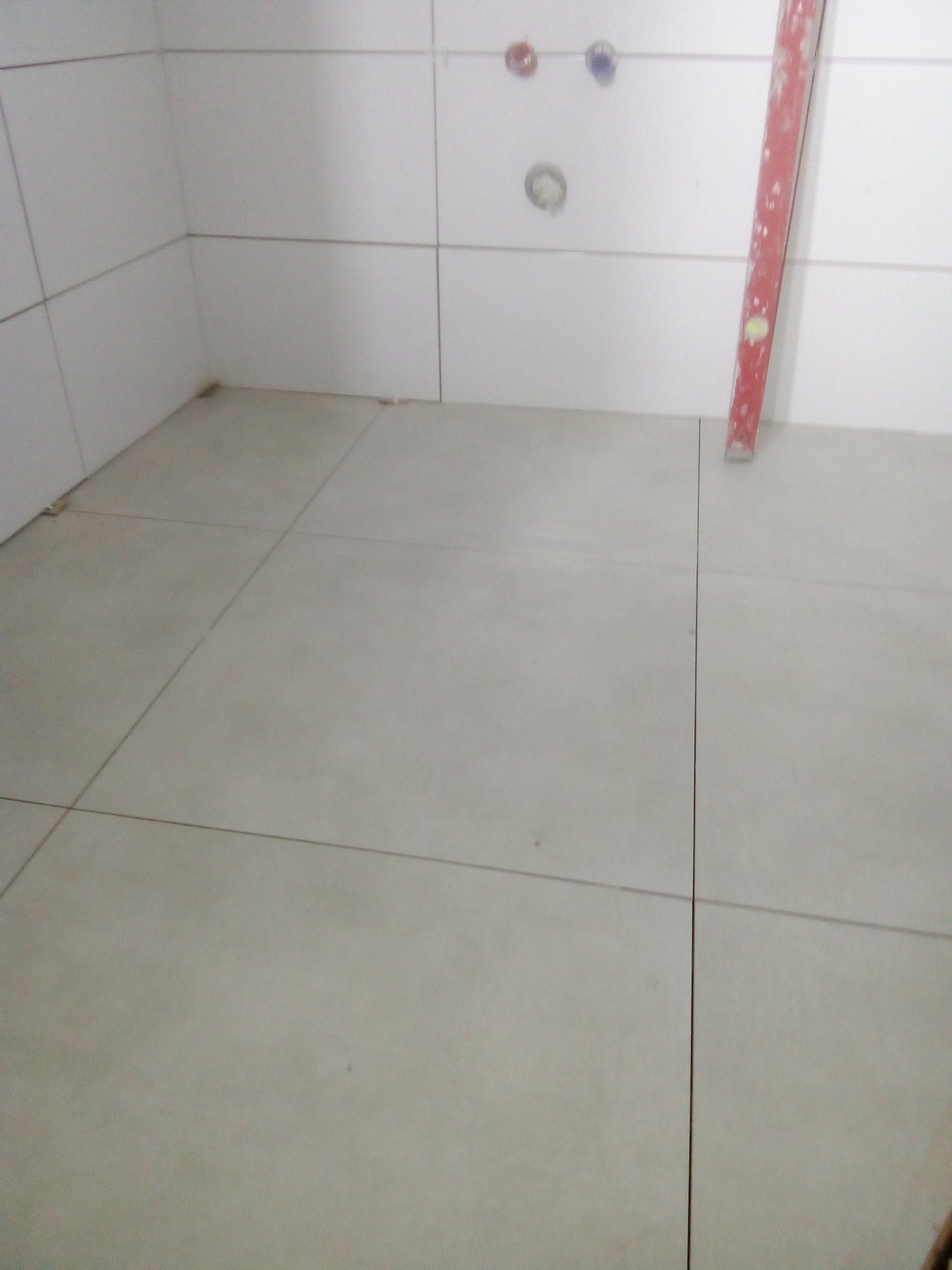 Świeżo ułożona, jasnoszara podłoga z płytek w rogu pomieszczenia, widoczna poziomica oparta o ścianę i wystające rury instalacji wodnej.