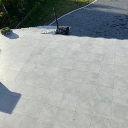 krzysztof - Jasnoszara, płytkowa posadzka tarasu z widocznymi schodami, fragmentami mebli ogrodowych i kominem, sfotografowana z góry w słoneczny dzień.