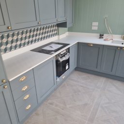 krzysztof - Nowoczesna kuchnia w odcieniach szarości i zieleni z geometrycznym wzorem na ścianie nad blatem, złotymi uchwytami i jasną podłogą ułożoną w jodełkę.