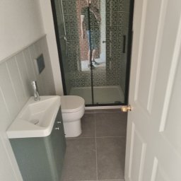 krzysztof - Nowoczesna łazienka z kabiną prysznicową z czarnymi ramami, wiszącą umywalką z zieloną szafką, szarą płytką podłogową i panelami ściennymi w pionowe paski, widziana zza uchylonych białych drzwi.