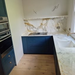 krzysztof - Niebieska zabudowa kuchenna z piekarnikiem i marmurowym blatem oraz backsplashiem z widocznymi przewodami elektrycznymi i gniazdkami przed wykończeniem.