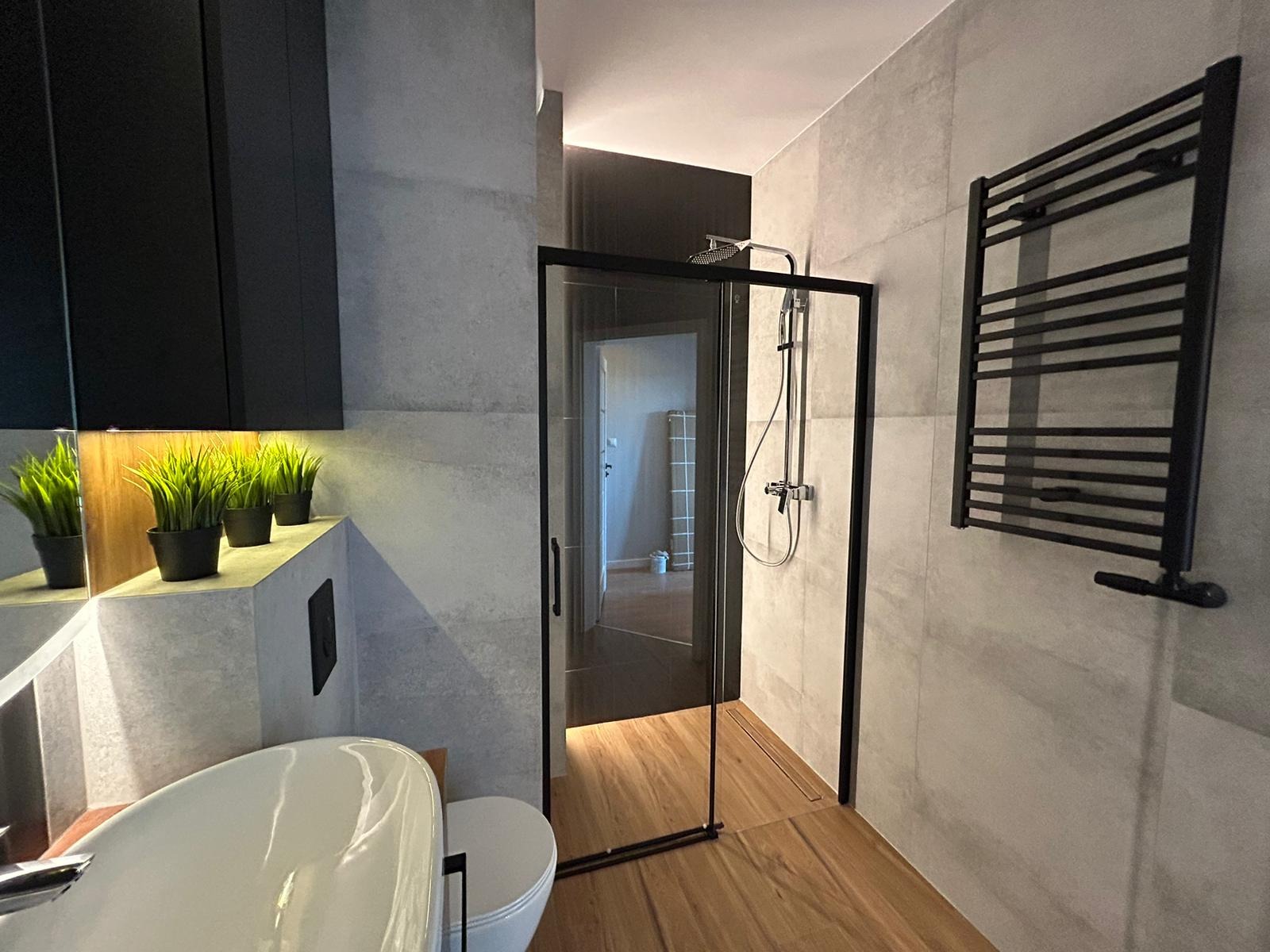 Nowoczesna łazienka z kabiną prysznicową typu walk-in z czarnymi ramami, grzejnikiem drabinkowym w kolorze czarnym, umywalką nablatową i drewnopodobną podłogą.