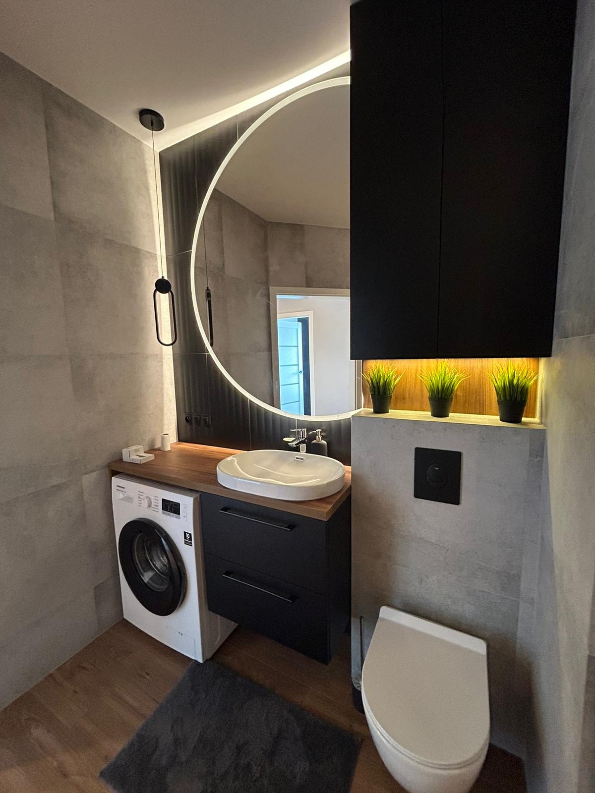 Nowoczesna łazienka w szarościach z drewnianym blatem, okrągłym lustrem podświetlanym LED, czarną szafką pod umywalką, pralką i wiszącą szafką z oświetleniem punktowym eksponującym dekoracyjne...