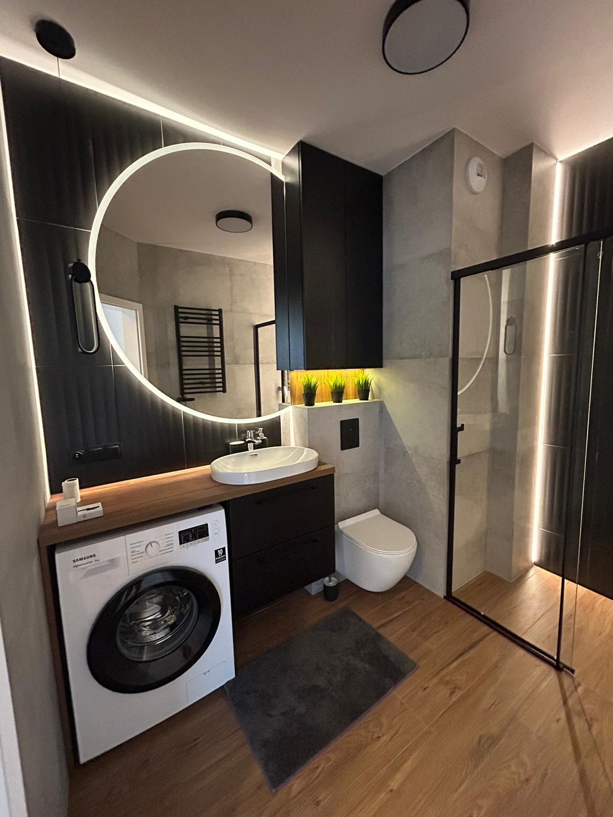 Nowoczesna łazienka z umywalką nablatową, podświetlanym lustrem w kształcie okręgu, pralką pod blatem i kabiną prysznicową walk-in z podświetleniem LED.