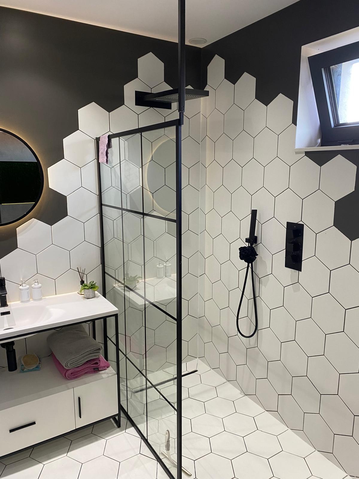Nowoczesna łazienka z heksagonalnymi płytkami w odcieniach bieli i szarości, czarną armaturą, kabiną prysznicową typu walk-in z czarnymi profilami i okrągłym lustrem z podświetleniem LED.