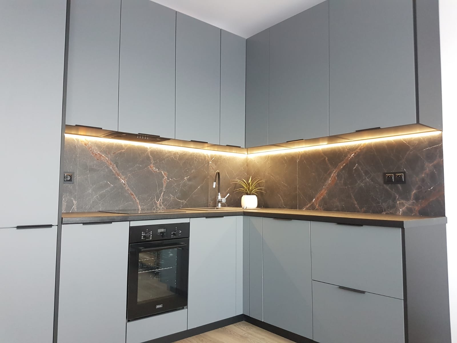 Nowoczesna kuchnia z szarymi szafkami, czarnym blatem i marmurowym panelem ściennym podświetlonym listwą LED. Na blacie minimalistyczna doniczka z rośliną.