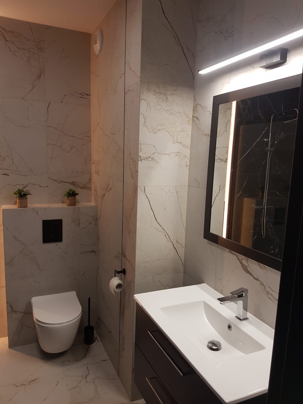 Nowoczesna łazienka wykończona płytkami imitującymi marmur, z wiszącą toaletą, czarną szczotką do WC, minimalistyczną umywalką z szafką i dużym lustrem z podświetleniem LED.