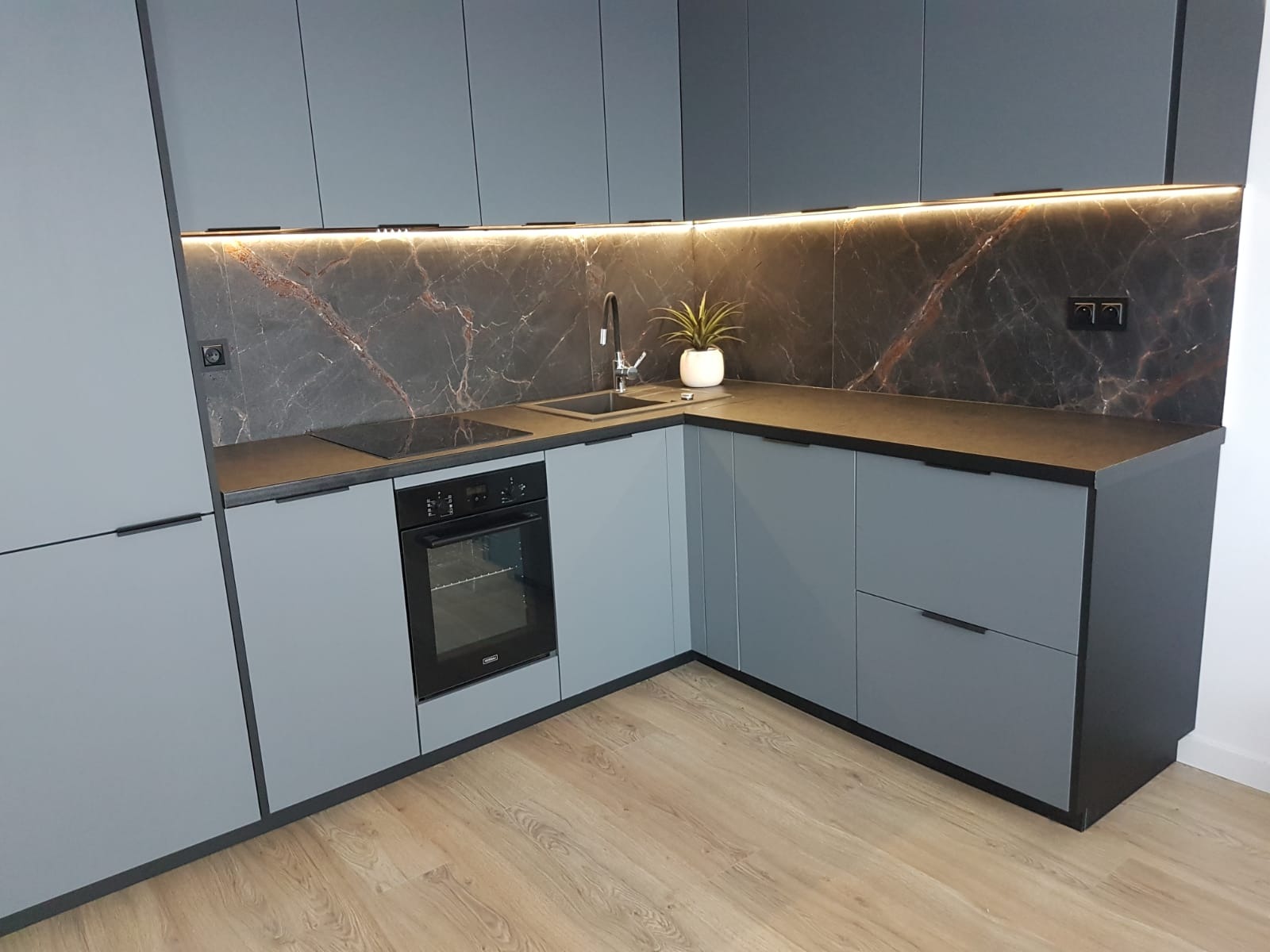 Nowoczesna kuchnia w odcieniach szarości z blatem z ciemnego kamienia, podświetleniem LED i marmurowym panelem nad blatem, uzupełniona o minimalistyczną armaturę i roślinę doniczkową.