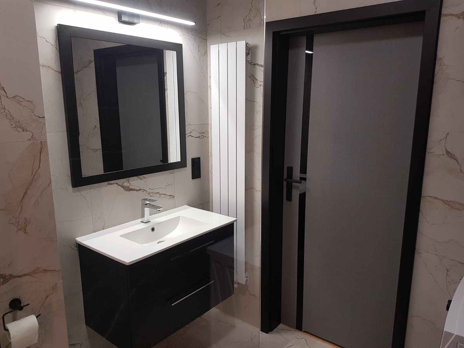 Nowoczesna łazienka z umywalką na szafce w kolorze czarnym, lustrem w czarnej ramie, białym grzejnikiem drabinkowym i drzwiami z szarą okleiną oraz czarną ościeżnicą, całość na tle marmurowych płytek.