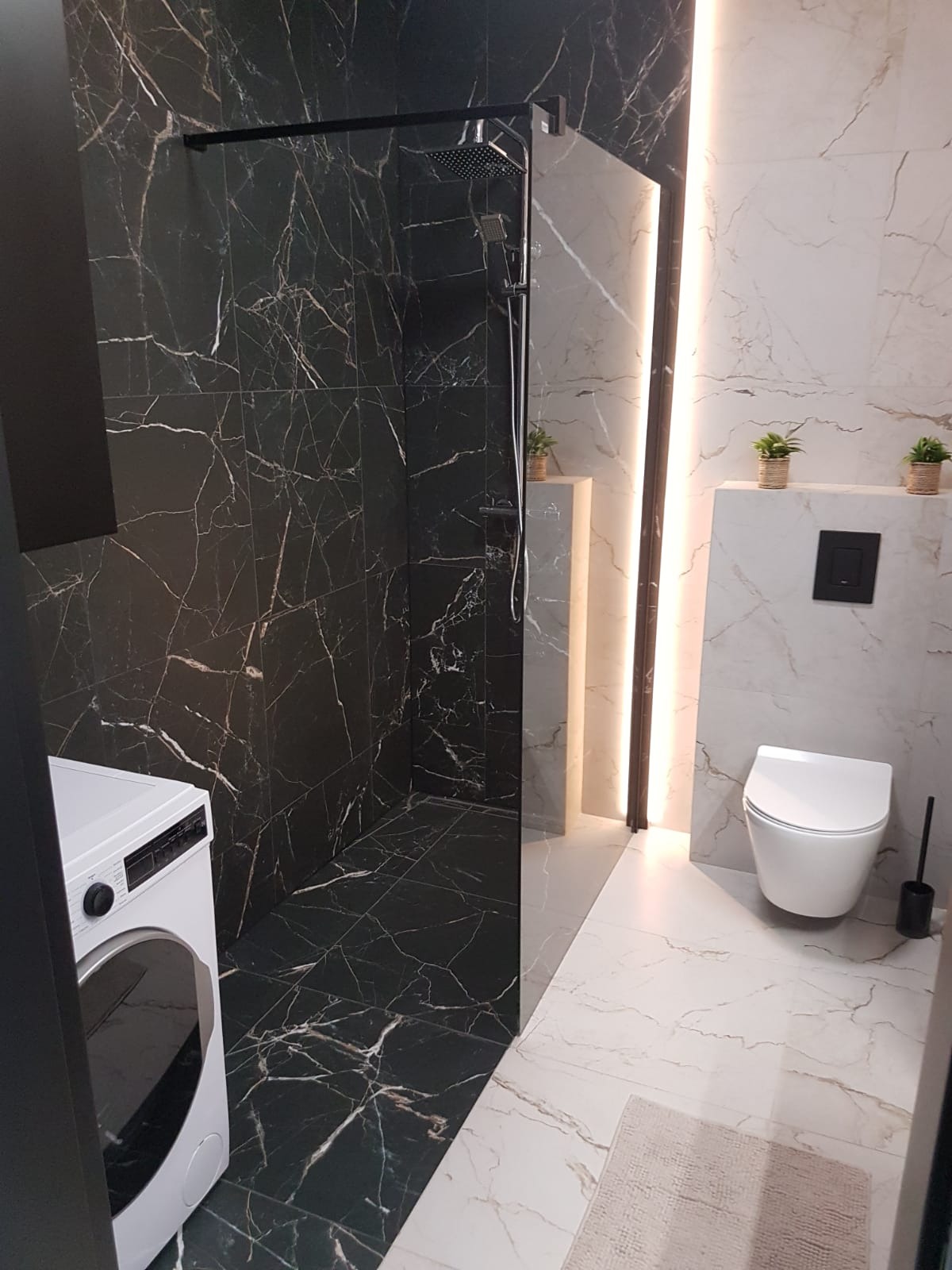 Nowoczesna łazienka z kabiną prysznicową typu walk-in wyłożoną czarnym marmurem z białymi żyłami, kontrastująca z jasnymi płytkami imitującymi marmur na pozostałej części ścian i podłogi; widoczna...