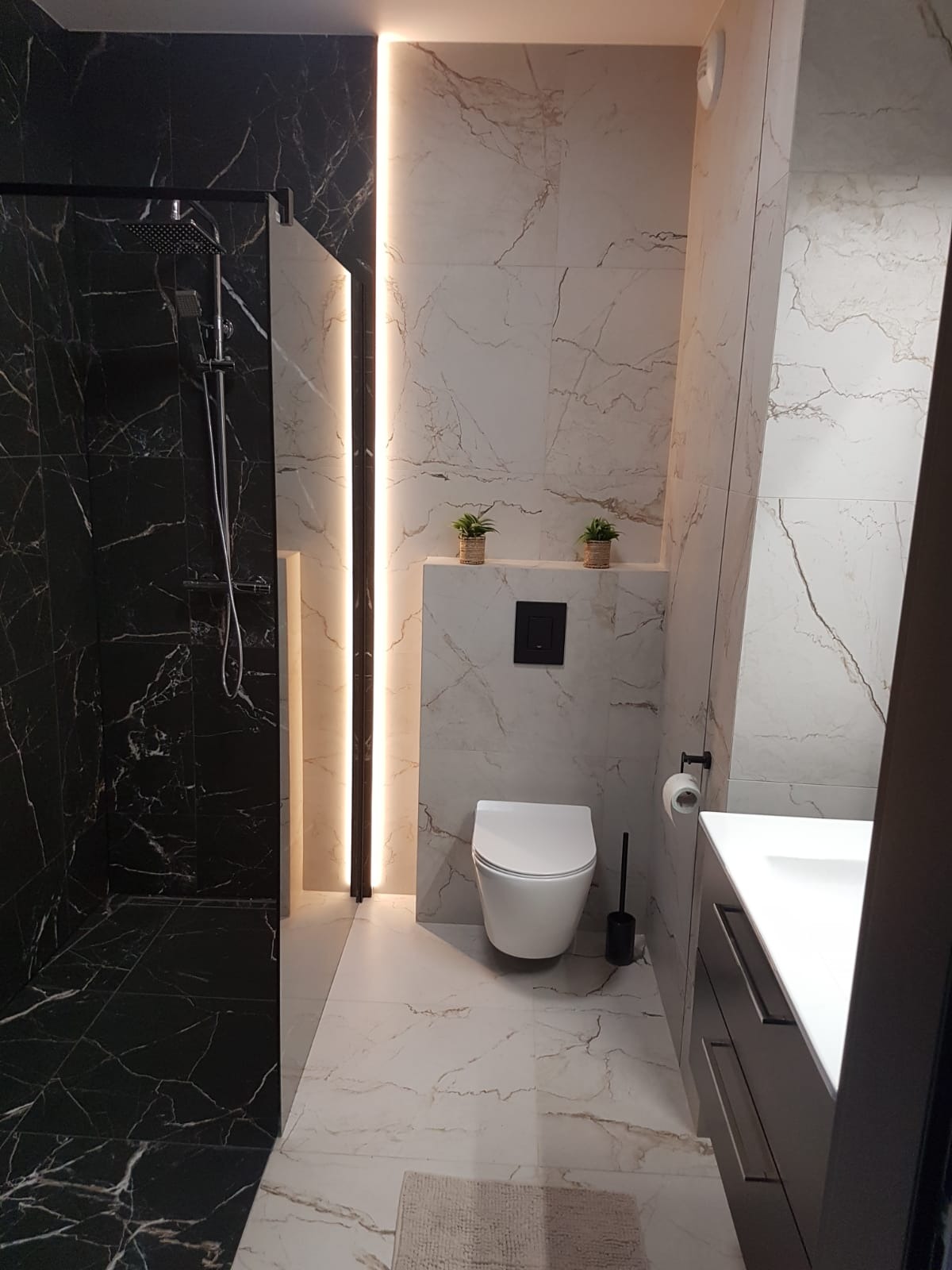 Nowoczesna łazienka z kabiną prysznicową wyłożoną czarnym marmurem z białymi żyłami, naprzeciwko której znajduje się toaleta podwieszana z czarnym przyciskiem spłukującym i umywalka z szufladami...