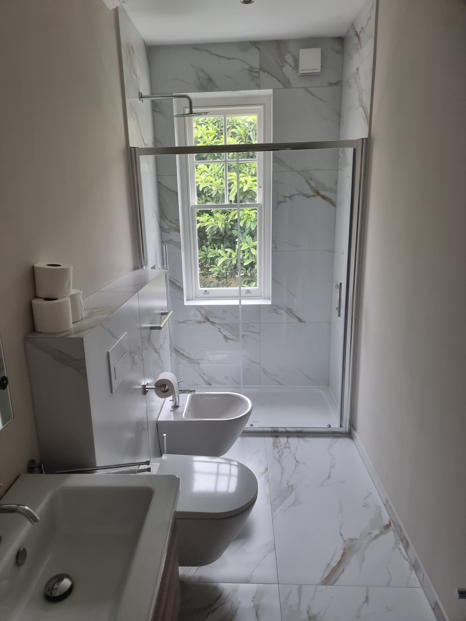 Nowoczesna łazienka z marmurowymi płytkami, kabiną prysznicową ze szklanymi drzwiami, wiszącą toaletą i bidetem, widok na okno z zielenią na zewnątrz, oraz rolki papieru toaletowego.