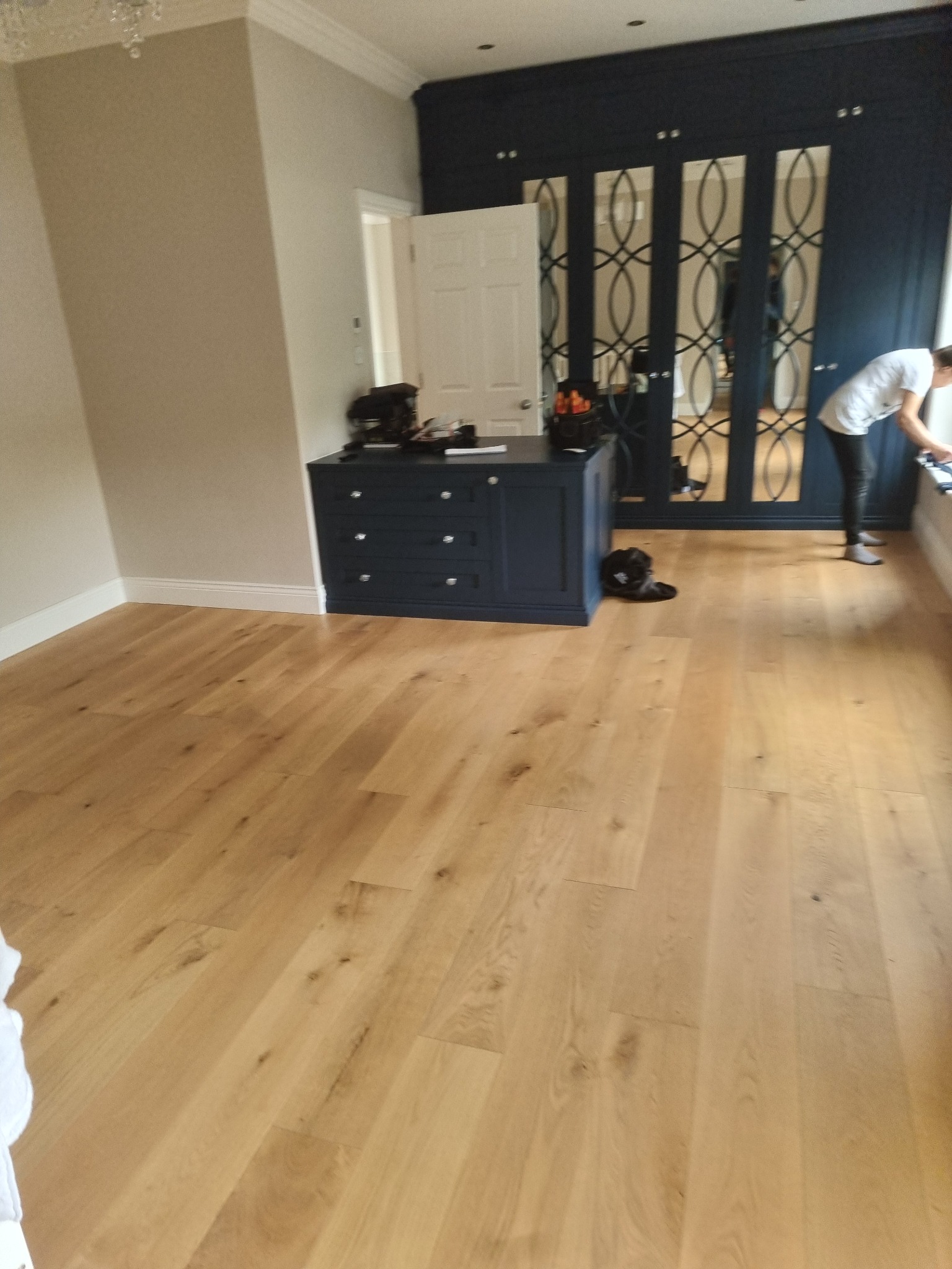 Jasna, dębowa podłoga w pokoju z granatową zabudową szafy z lustrami w ozdobnych ramach oraz komodą w tym samym kolorze; osoba czyści okno w tle.