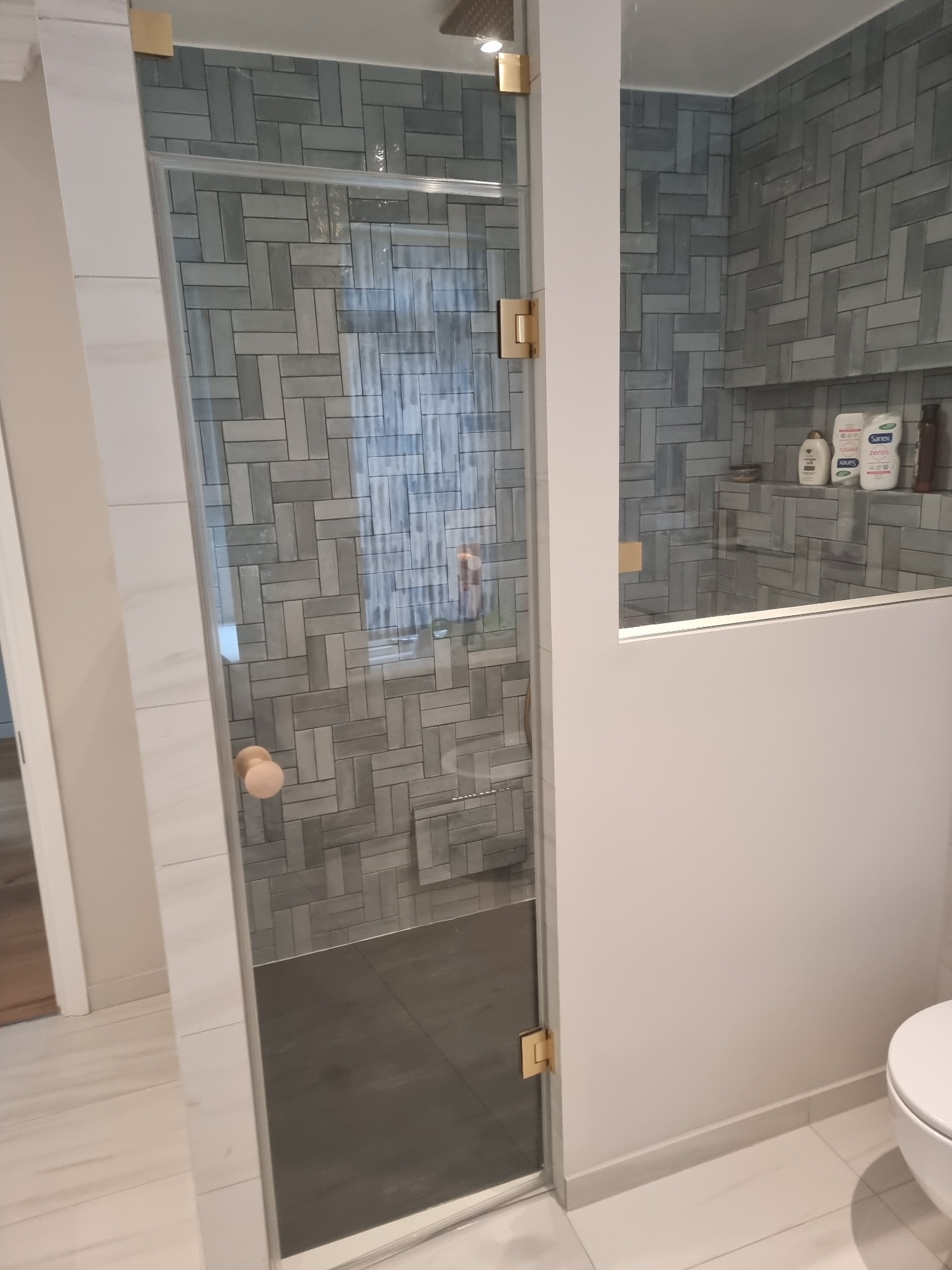 Nowoczesna kabina prysznicowa z szarymi płytkami ułożonymi w jodełkę, złotymi okuciami i drewnianą gałką, widok na półkę z kosmetykami.