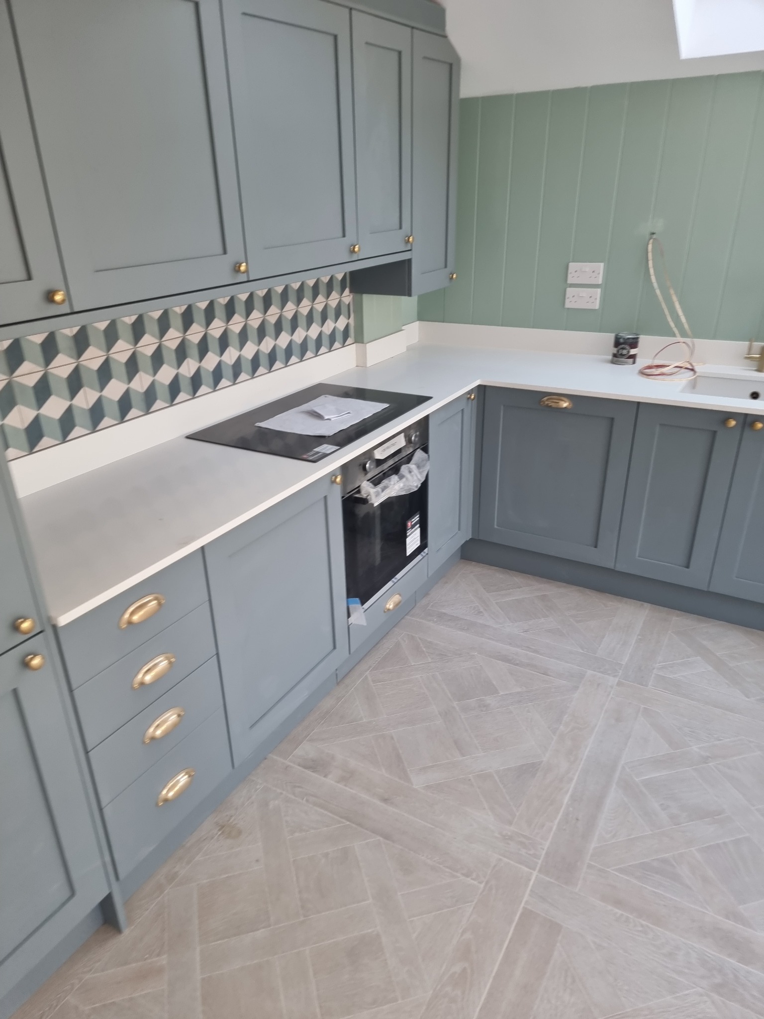 Nowoczesna kuchnia w odcieniach szarości i zieleni z geometrycznym wzorem na ścianie nad blatem, złotymi uchwytami i jasną podłogą ułożoną w jodełkę.