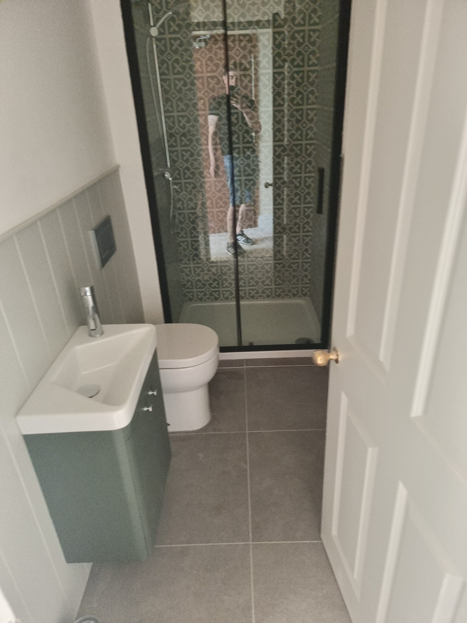 Nowoczesna łazienka z kabiną prysznicową z czarnymi ramami, wiszącą umywalką z zieloną szafką, szarą płytką podłogową i panelami ściennymi w pionowe paski, widziana zza uchylonych białych drzwi.