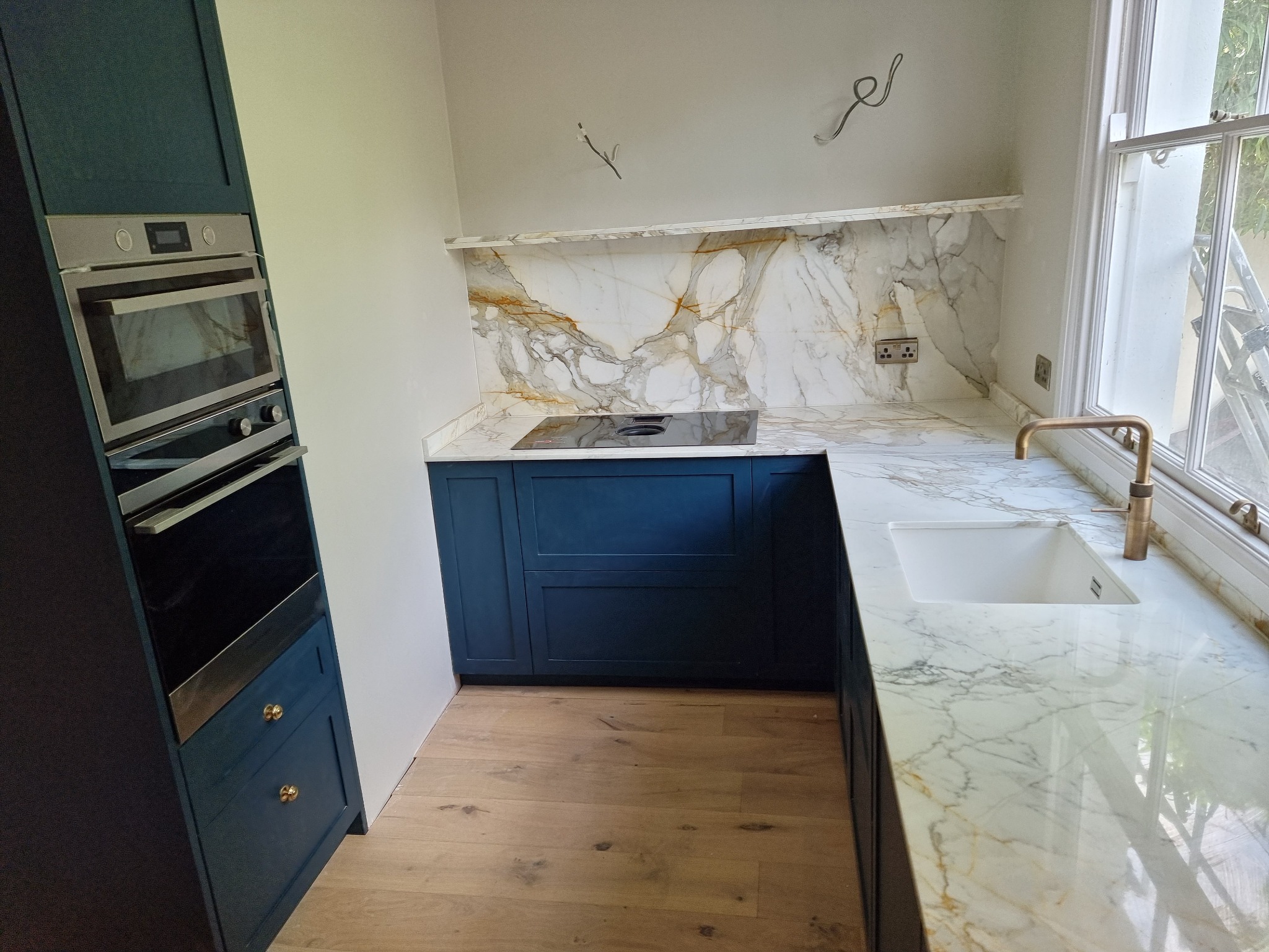 Niebieska zabudowa kuchenna z piekarnikiem i marmurowym blatem oraz backsplashiem z widocznymi przewodami elektrycznymi i gniazdkami przed wykończeniem.