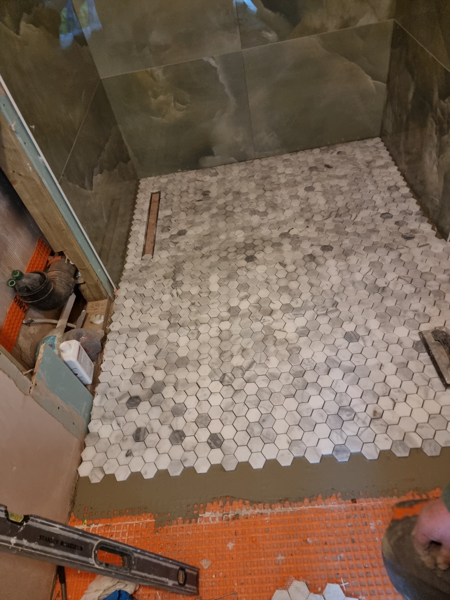 Układanie mozaiki heksagonalnej w szaro-białych odcieniach na podłodze prysznica, widoczny odpływ liniowy, poziomica i resztki zaprawy klejowej na pomarańczowej macie hydroizolacyjnej.