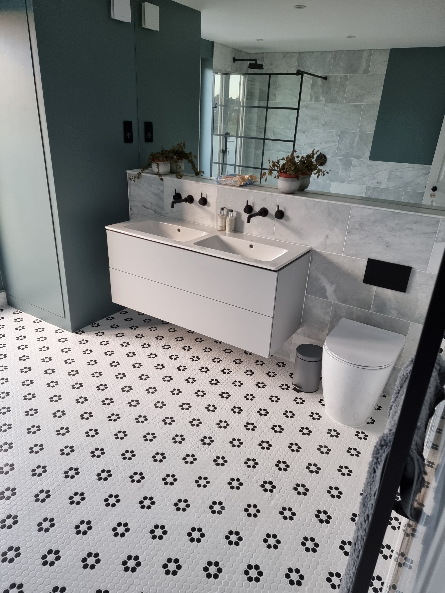 Nowoczesna łazienka z białą, podwieszaną szafką z dwiema umywalkami i czarną armaturą, mozaikową podłogą w biało-czarne kwiatki, szarą ścianą z marmurową wstawką i kabiną prysznicową ze szklaną...