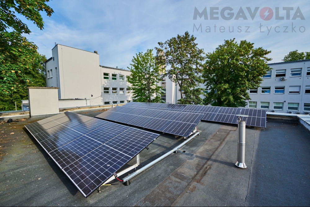 Instalacja paneli słonecznych na płaskim dachu budynku, widoczna szara elewacja i zieleń drzew, logo firmy w prawym górnym rogu.