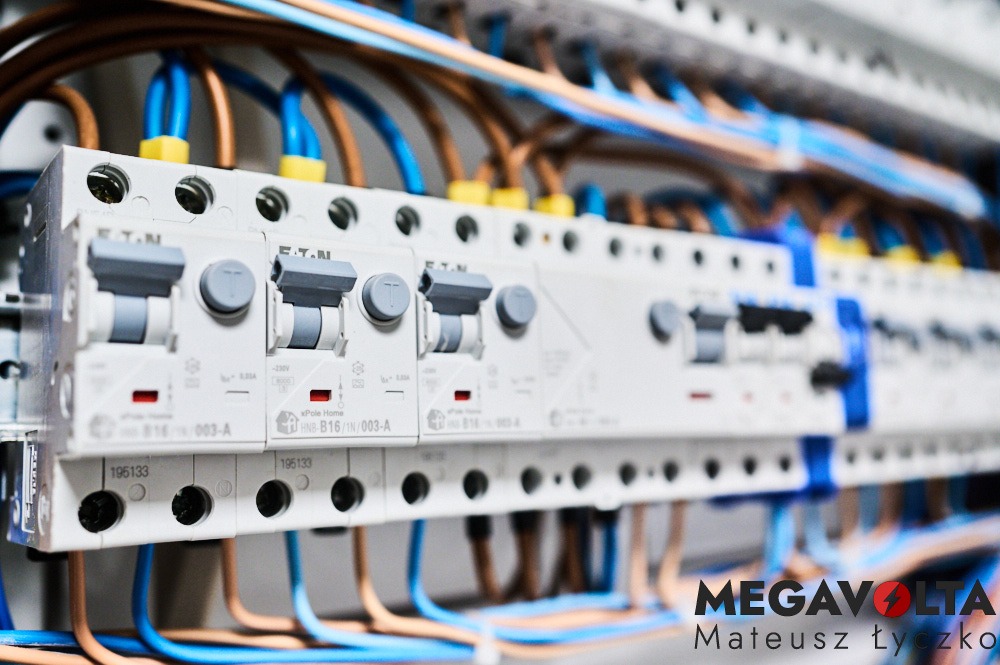 Szczegółowe ujęcie tablicy rozdzielczej z zamontowanymi bezpiecznikami EATON, widoczne niebieskie i brązowe przewody podłączone do aparatury, logo firmy MEGAVOLTA w prawym dolnym rogu.