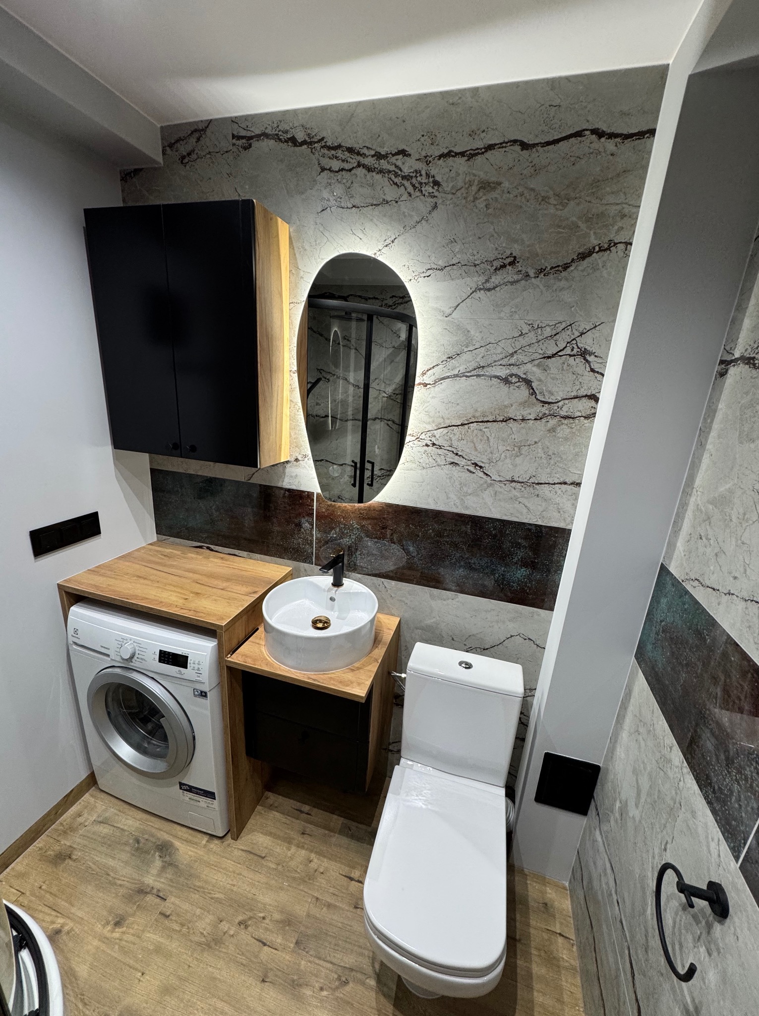 Nowoczesna łazienka z umywalką nablatową, podświetlanym owalnym lustrem, pralką zabudowaną pod blatem, wiszącą szafką i toaletą, wykończona płytkami imitującymi marmur z ciemnymi wstawkami.