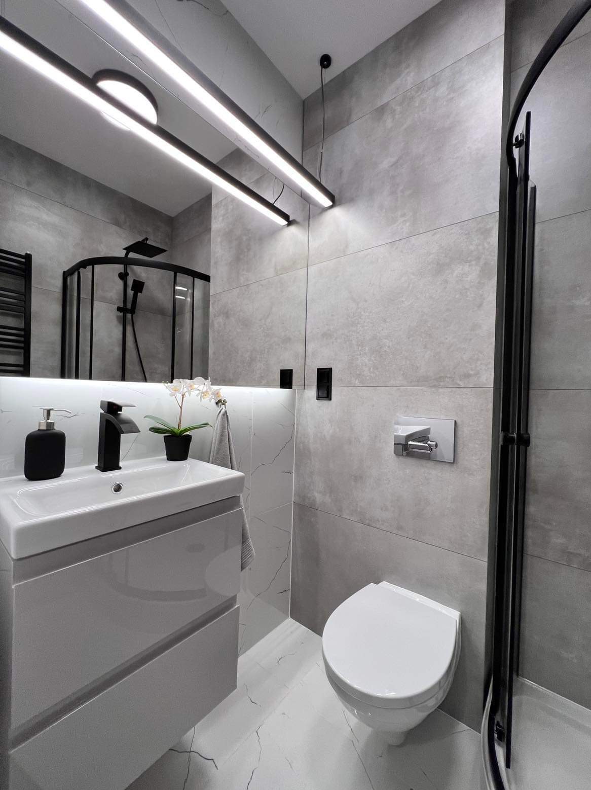 Nowoczesna łazienka z szarymi płytkami imitującymi beton, białymi marmurowymi płytkami na podłodze, czarną armaturą i kabiną prysznicową z czarnymi ramami, oświetlona podłużnymi lampami LED.