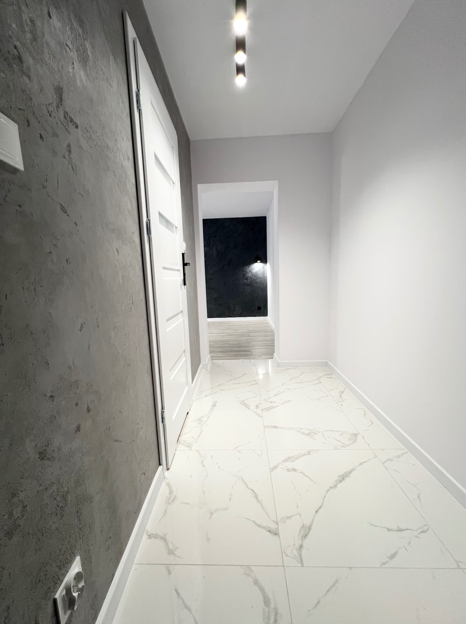 Nowoczesny korytarz z szarą ścianą o fakturze betonu, białymi drzwiami i podłogą z płytek imitujących marmur, oświetlony listwą z reflektorami.