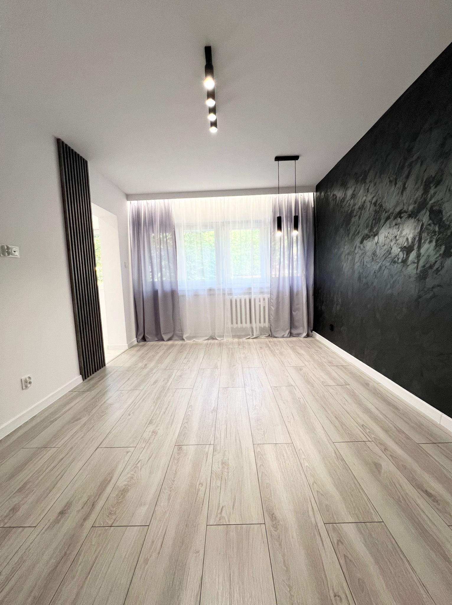 Jasne panele podłogowe ułożone w przestronnym, nowocześnie wykończonym pokoju z czarną ścianą o fakturze betonu, białymi ścianami, zasłonami w oknie i minimalistycznym oświetleniem sufitowym.