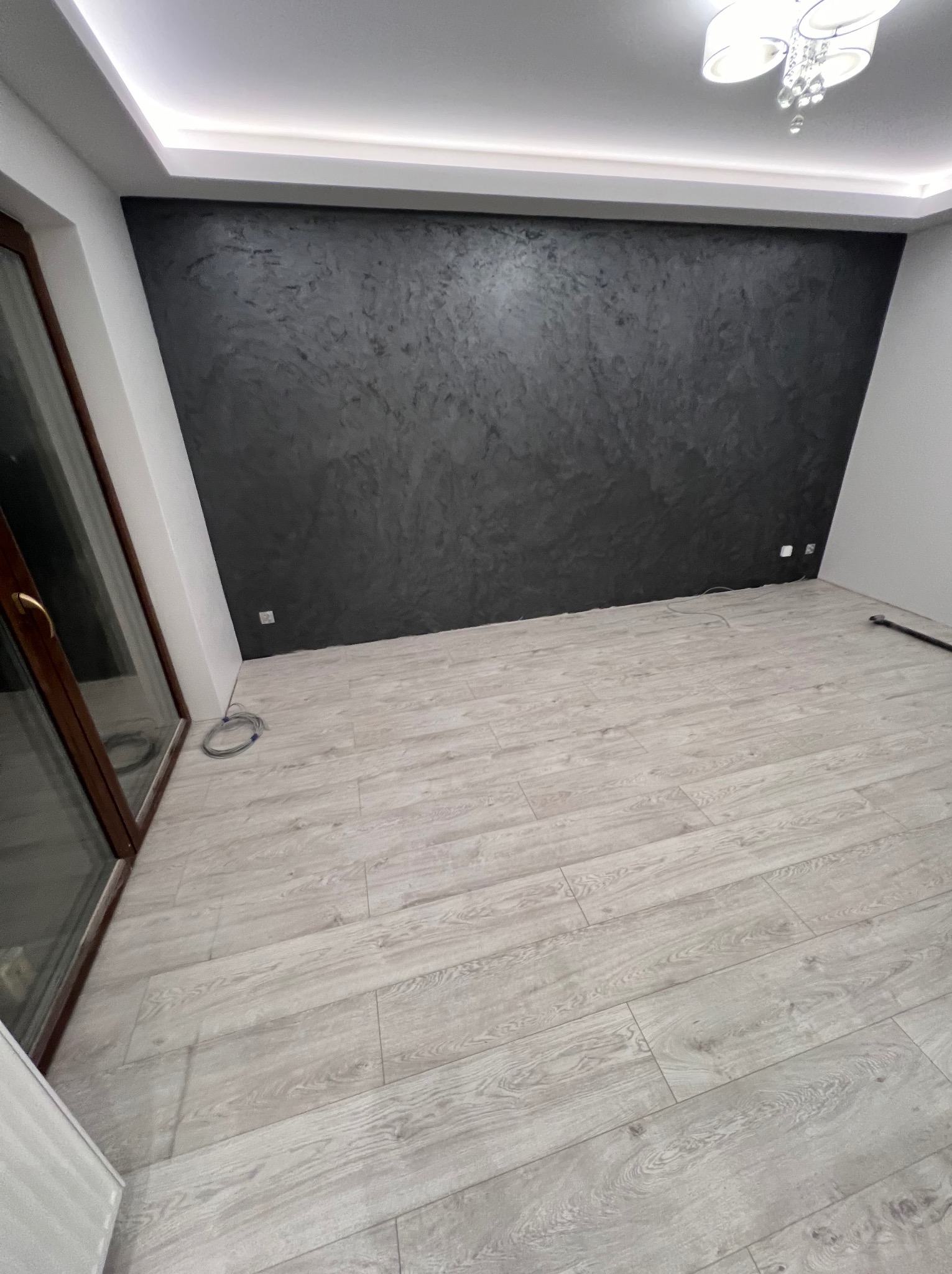 Wykończone wnętrze pokoju z szarą ścianą o fakturze betonu, jasnymi panelami podłogowymi ułożonymi w jodełkę i białym sufitem z oświetleniem LED, widoczne gniazdka elektryczne.