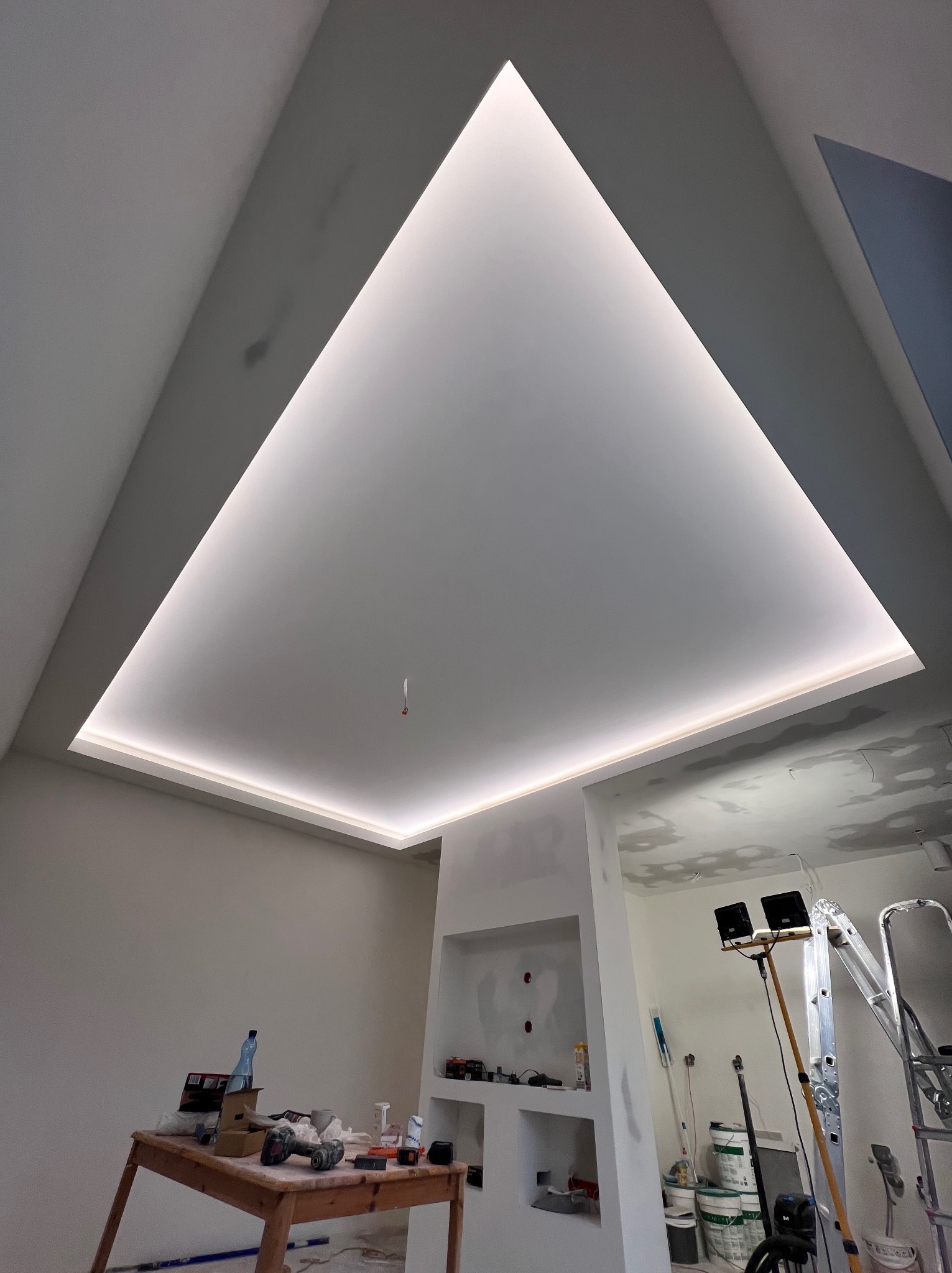 Sufit podwieszany z trójkątnym oświetleniem LED, widoczne narzędzia na stole, drabina i lampa budowlana, ściany przygotowane do malowania, wnęka w ścianie.