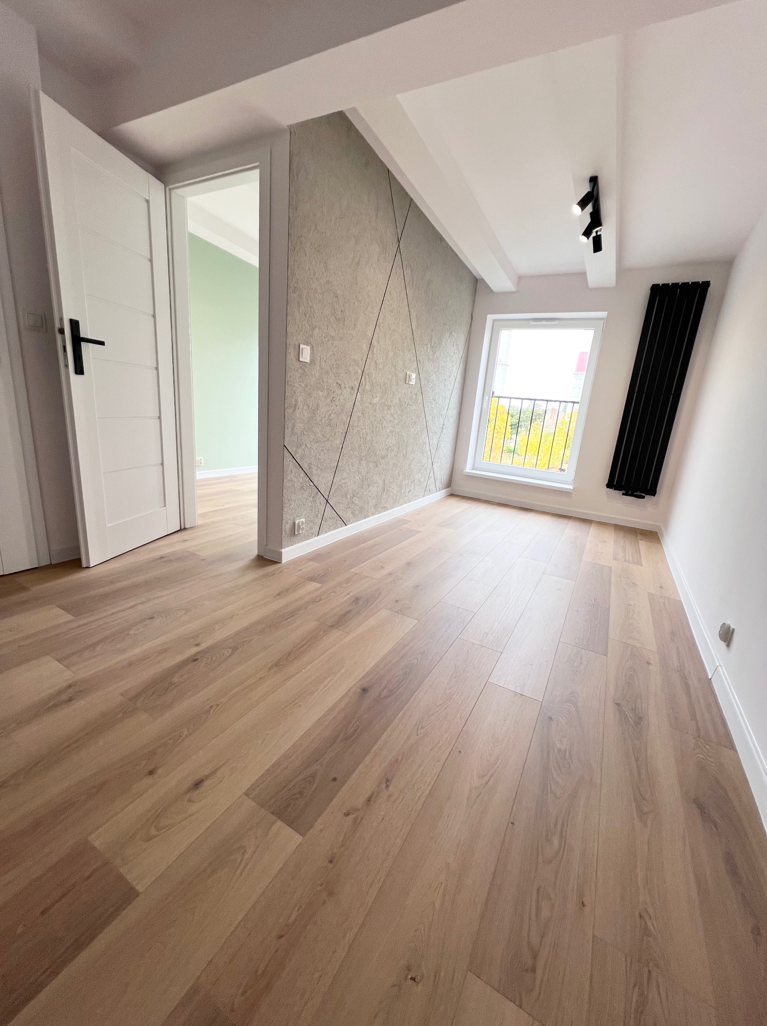 Jasne panele podłogowe w pokoju z oknem dachowym, szarą ścianą z geometrycznym wzorem i czarnym grzejnikiem żeberkowym.