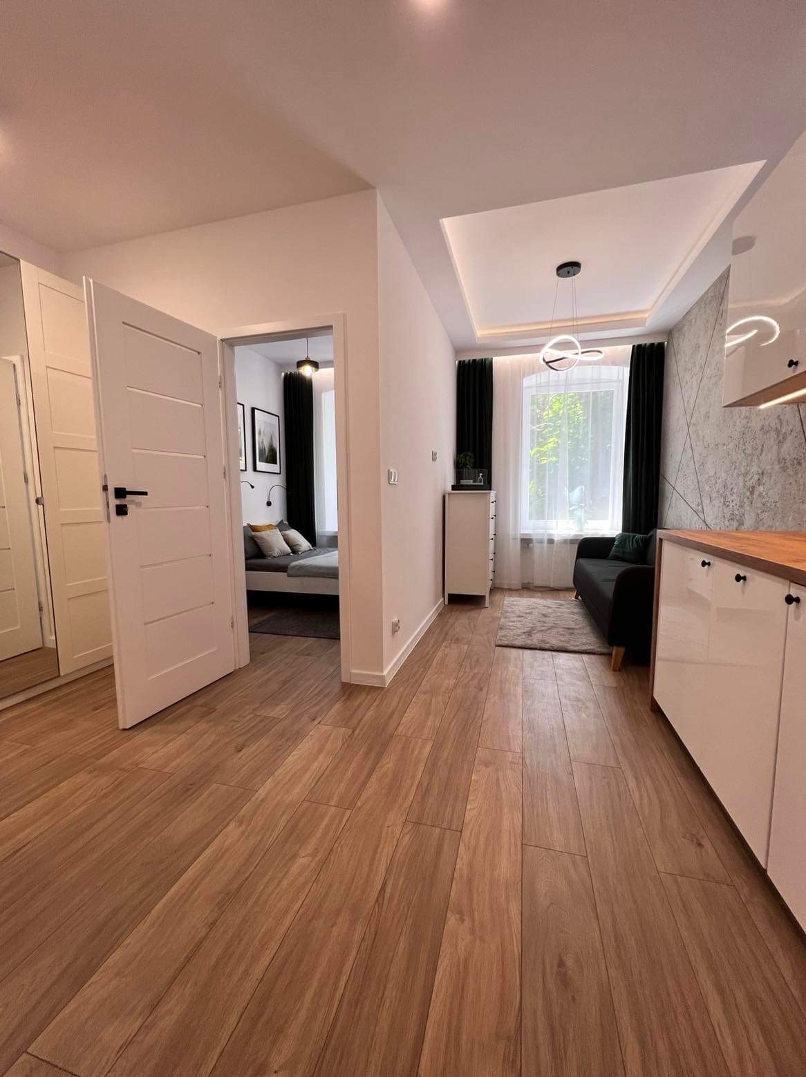 Jasne, przestronne wnętrze mieszkania po remoncie z drewnopodobną podłogą, otwartą sypialnią i nowoczesnym oświetleniem.