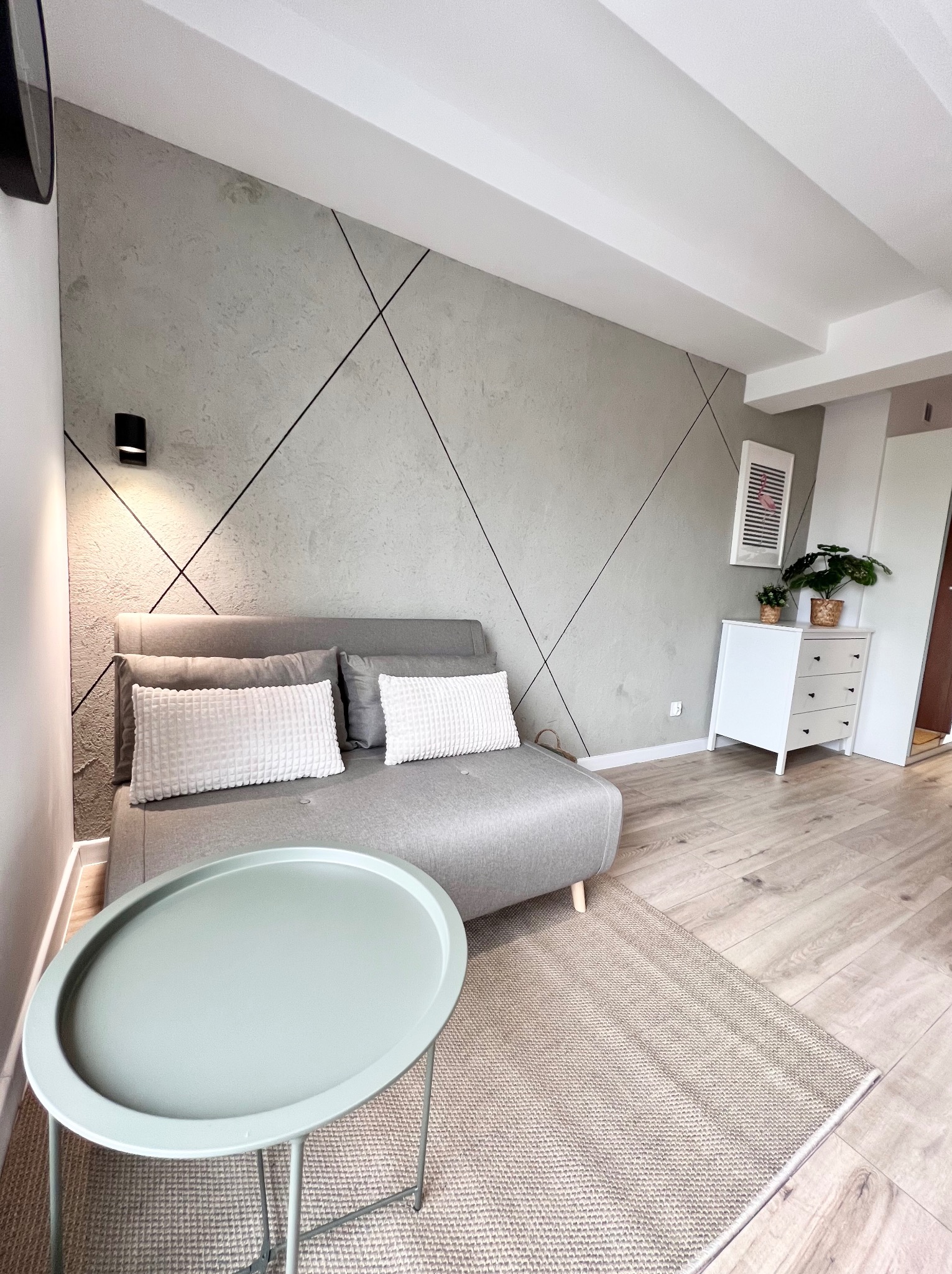 Szara sofa z białymi poduszkami na tle ściany z dekoracyjną fakturą i czarnymi liniami, jasne panele podłogowe, okrągły stolik w kolorze miętowym, biała komoda z roślinami, nowoczesne wnętrze...