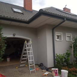 Malowanie mieszkan Vitalii Kalynchuk - Dom jednorodzinny po malowaniu elewacji, widoczna drabina, pędzle i farby. Świeżo pomalowana ściana w odcieniach szarości. Dachówka ceramiczna i ceglany komin.