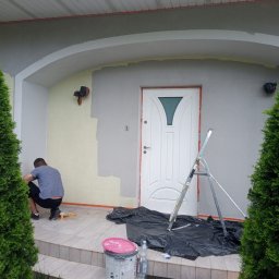 Malowanie mieszkan Vitalii Kalynchuk - Malowanie elewacji domu: mężczyzna w trakcie pracy, widoczne pędzle, drabina, farba i zabezpieczona folią strefa malowania. Częściowo pomalowana ściana.