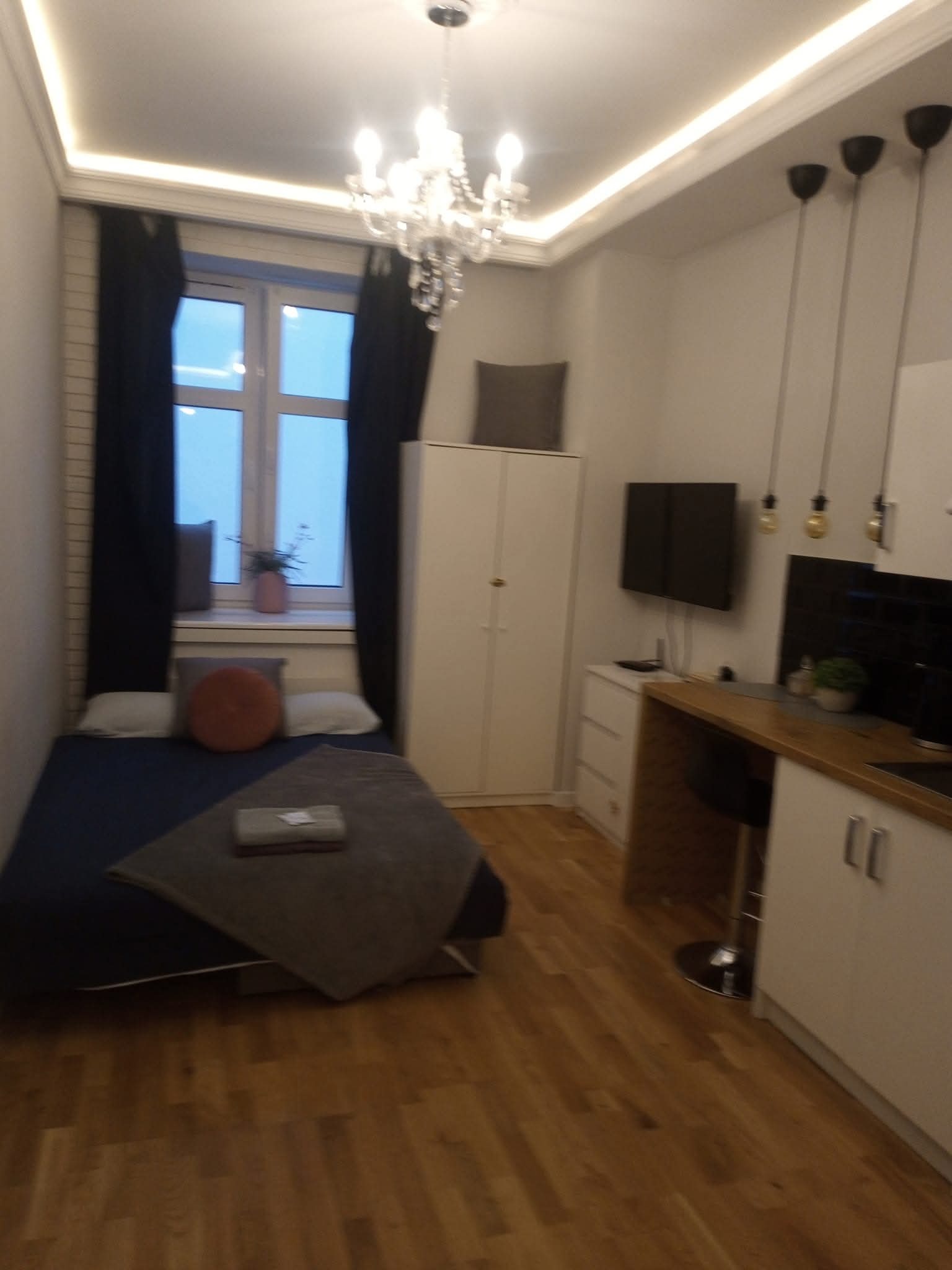 Nowoczesne studio: drewniana podłoga, aneks kuchenny, łóżko przy oknie z czarnymi zasłonami, oświetlenie sufitowe LED i żyrandol. Minimalistyczny design.